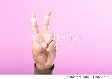 Hand with bandages or plaster on the index and middle fingers forming a 'V' sign, symbolizing resilience and the ability to keep working and going despite minor injuries Hand with bandages or plaster on the index and middle fingers forming a 'V' sign, symbolizing resilience and the ability to keep working and going despite minor injuries 120633426