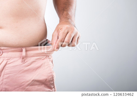 Man with a larger physique stands adjusting the waistband of his pants to check if they are tight or loose, reflecting the importance of monitoring body shape and overall health for men 120633462