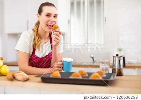 Portrait of cheerful woman in apron eating just cooked cupcakes 120636259