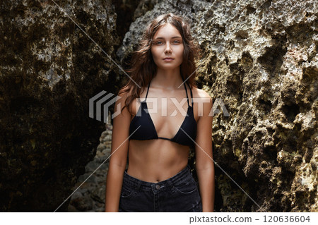 Beautiful young woman in bikini standing by a rock 120636604