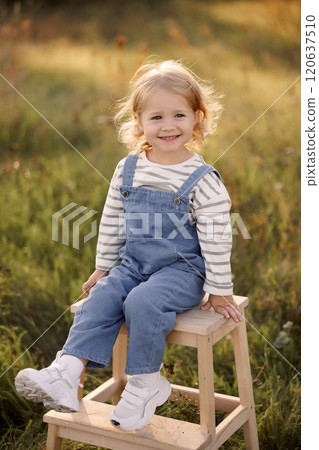 Little smiling child in denim overalls sits on a wooden stool in the field.  120637510