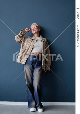 Vertical full length portrait of modern senior woman with white hair wearing glasses and casual outfit with baggy jeans posing against blue wall in studio 120639120