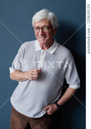 Vertical studio portrait of white haired senior man smiling at camera posing casually against blue background in studio and wearing glasses 120639129