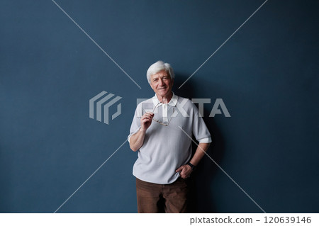 Minimal front view portrait of smiling senior man looking at camera standing against blue wall in studio with natural light and holding glasses in hand, copy space 120639146