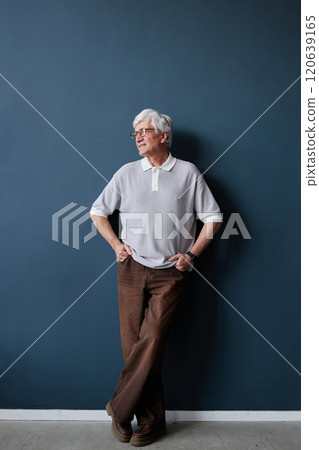 Vertical full length portrait of stylish senior man with white hair looking away while posing relaxed against blue wall in studio with soft natural light 120639165