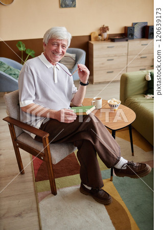 Vertical full length portrait of retired senior man with while hair looking at camera smiling cheerfully in cozy home and reading book 120639173