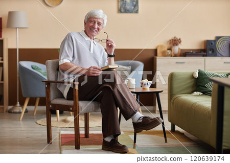 Full length portrait of joyful senior man with white hair smiling at camera in cozy home while reading book sitting on chair, copy space 120639174