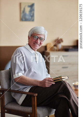 Portrait of Caucasian senior man with grey hair reading book sitting on chair in cozy home and looking at camera smiling 120639175