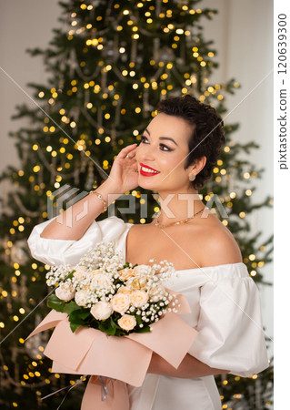 A woman stands in front of a Christmas tree, holding a bouquet A woman stands in front of a Christmas tree, holding a bouquet 120639300