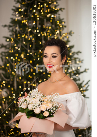 A beautiful woman with a bouquet of roses stands near a Christmas tree decorated with lights and tinsel 120639302