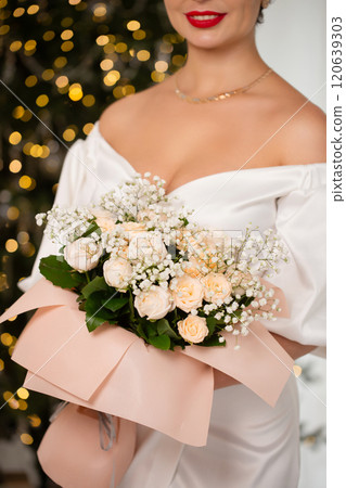 A woman stands in front of a Christmas tree, holding a bouquet of flowers, close-up A woman stands in front of a Christmas tree, holding a bouquet of flowers, close-up 120639303
