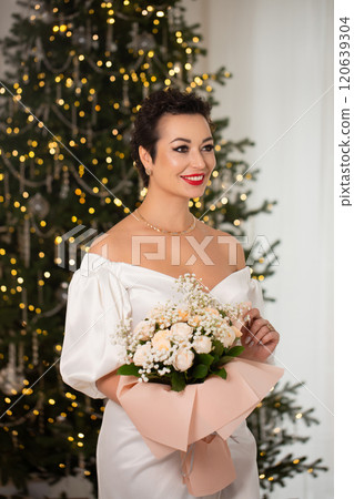 A woman with a bouquet of roses stands near a Christmas tree decorated with lights and tinsel 120639304