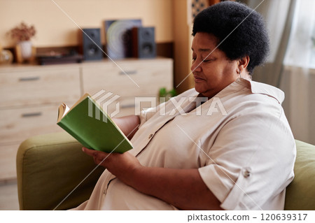 Side view portrait of African American senior woman reading book sitting on couch and enjoying relaxing day at home copy space Side view portrait of African American senior woman reading book sitting on couch and enjoying relaxing day at home copy space 120639317