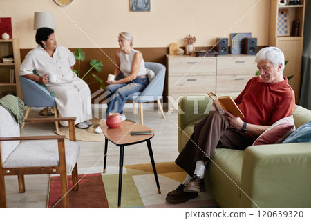 Cozy scene with family enjoying quiet day at home and white haired senior man reading book while sitting on couch in foreground copy space 120639320