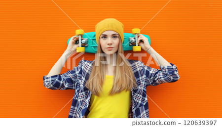 Portrait of stylish young girl with skateboard in hat, modern teenage female posing on orange 120639397