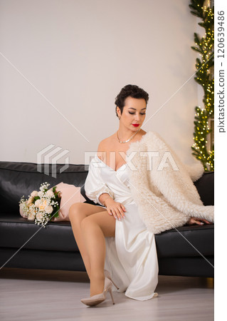 A woman in a white dress with a bouquet of roses sits on a black couch. She is wearing a white fur coat and a necklace 120639486