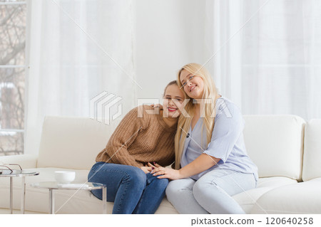 Two women are sitting on a white couch, one of them is wearing a brown sweater Two women are sitting on a white couch, one of them is wearing a brown sweater 120640258