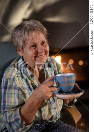 Elderly woman sitting near a fireplace, smiling warmly while holding a blue teacup, enjoying a cozy and relaxed evening. Elderly woman sitting near a fireplace, smiling warmly while holding a blue teacup, enjoying a cozy and relaxed evening. 120640343