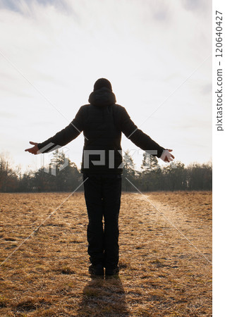 A man stands in a field with his arms outstretched, looking up at the sky 120640427