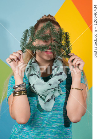 A woman covered her face with Christmas tree branches and smiles. Behind her is a colorful background A woman covered her face with Christmas tree branches and smiles. Behind her is a colorful background 120640434