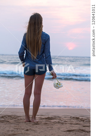 A woman stands on the beach holding a white plumeria flower . The sky is pink and the sun is setting 120641011