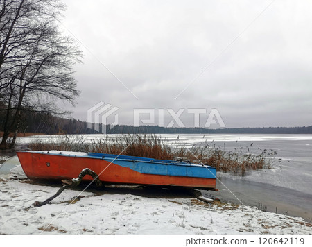 An old boat on the shore of a frozen pond - lake against the background of a forest in the snow. Ice on the lake, ice rink and winter fishing An old boat on the shore of a frozen pond - lake against the background of a forest in the snow. Ice on the lake, ice rink and winter fishing 120642119