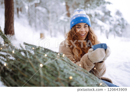 Young woman drinks a hot drink from a thermos while sitting in a snowy forest with a Christmas tree. 120643909