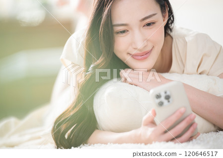 A young woman operating a smartphone in bed or in the living room after waking up A young woman operating a smartphone in bed or in the living room after waking up 120646517