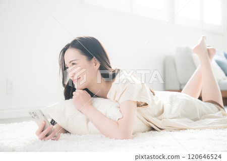 一位微笑的年輕女性醒來後在床上或客廳裡拿著智慧型手機思考和抬頭的全身廣角 一位微笑的年輕女性醒來後在床上或客廳裡拿著智慧型手機思考和抬頭的全身廣角 120646524