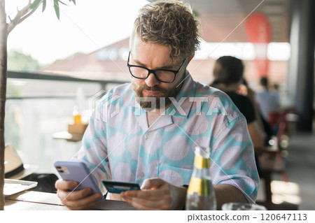Millennial man with glasses and a patterned shirt uses his smartphone and credit card while sitting at an outdoor cafe. Modern technology online shopping and digital payments concept Millennial man with glasses and a patterned shirt uses his smartphone and credit card while sitting at an outdoor cafe. Modern technology online shopping and digital payments concept 120647113