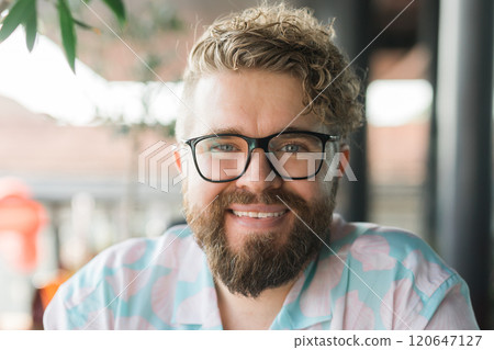 Close-up portrait of a man with glasses, curly hair, and beard, sitting outdoors at a cafe. Neutral expression and natural lighting highlight individuality and a calm, thoughtful demeanor. Millennial 120647127