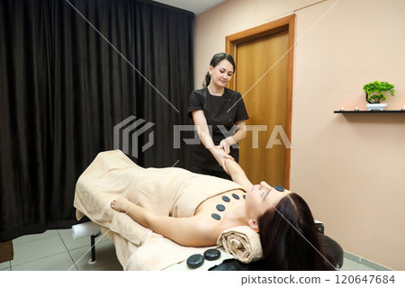 A Skilled Female Massage Therapist Gently Massaging a Client's Hands with Warm Basalt Stones, Creating a Tranquil Atmosphere That Promotes Relaxation and Well-Being, While Highlighting the Therapeutic A Skilled Female Massage Therapist Gently Massaging a Client's Hands with Warm Basalt Stones, Creating a Tranquil Atmosphere That Promotes Relaxation and Well-Being, While Highlighting the Therapeutic 120647684