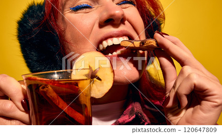 Closed-up portrait of young red haired woman biting gingerbread cookie and drinking mulled wine against yellow background Closed-up portrait of young red haired woman biting gingerbread cookie and drinking mulled wine against yellow background 120647809
