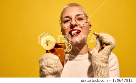 Smiling young woman with short hair, in knitted gloves eating gingerbread cookies and drinking hot cider, against bright yellow background Smiling young woman with short hair, in knitted gloves eating gingerbread cookies and drinking hot cider, against bright yellow background 120647811