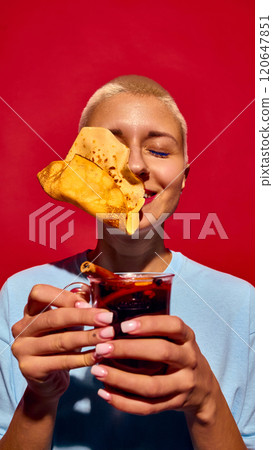 Funny image of young woman with short hair, holding glass of mulled wine with crepe pancake flying to her face, set against red background Funny image of young woman with short hair, holding glass of mulled wine with crepe pancake flying to her face, set against red background 120647851