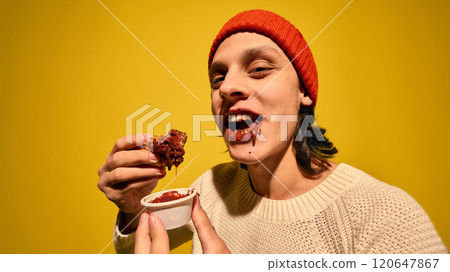 Young man in red beanie hat, with mouth in chocolate, eating churros with chocolate, set against bright yellow background 120647867
