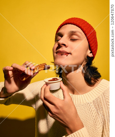 Young man in red beanie hat, eating churros with chocolate, set against bright yellow background. Enjoying sweet food Young man in red beanie hat, eating churros with chocolate, set against bright yellow background. Enjoying sweet food 120647870