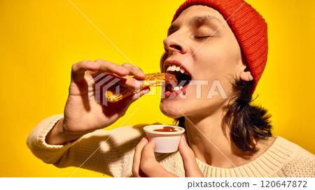 Young man in red beanie hat, eating churros with chocolate, set against bright yellow background. Enjoying sweet food Young man in red beanie hat, eating churros with chocolate, set against bright yellow background. Enjoying sweet food 120647872