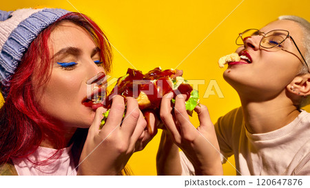 Two women emotional and passionately eating hot dog with ketchup and lettuce, against bright yellow background. Street food popular snack Two women emotional and passionately eating hot dog with ketchup and lettuce, against bright yellow background. Street food popular snack 120647876