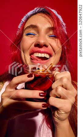 Close-up of smiling woman with red hair enjoying glass of mulled wine, set against vibrant red background. 120647922