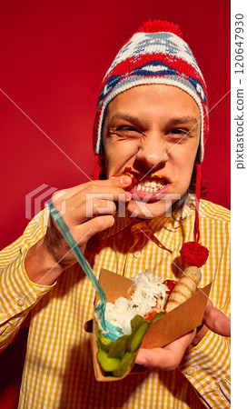 Delicious bite. Young man in yellow sweater and hat, emotionally eating grilled sausage with sauerkraut and pickles against bright red background 120647930