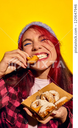 Close-up portrait of cheerful young redhead woman in hat and checkered shower, emotionally eating sugar powdered donuts against bright yellow background Close-up portrait of cheerful young redhead woman in hat and checkered shower, emotionally eating sugar powdered donuts against bright yellow background 120647946