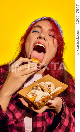 Delicious lick. Young redhead woman enjoying delicious sweet mini donuts with powdered sugar, eating with passion against vivid yellow background Delicious lick. Young redhead woman enjoying delicious sweet mini donuts with powdered sugar, eating with passion against vivid yellow background 120647947