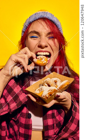 Close-up of emotionally young redhead woman in hat and checkered shirt, biting sweet powdered mini donuts again bright yellow background. Close-up of emotionally young redhead woman in hat and checkered shirt, biting sweet powdered mini donuts again bright yellow background. 120647948
