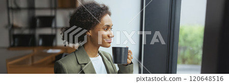 A woman in formal wear sips coffee, gazes out window 120648516