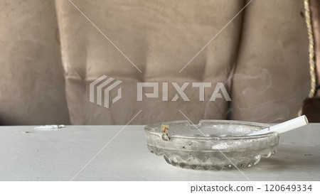 Cigarette in ashtray smoldering on a table near a couch 120649334