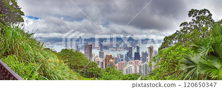 Panoramic daytime view of Hong Kong skyline, surrounded by lush green hills and a busy harbor 120650347
