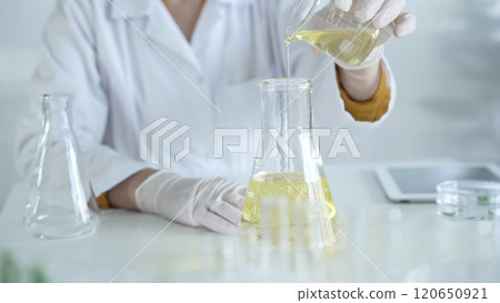 A scientist, wearing a lab coat and white protective gloves, is pouring a yellow oily liquid from one beaker to another in laboratory, close up. Medicine and science concept A scientist, wearing a lab coat and white protective gloves, is pouring a yellow oily liquid from one beaker to another in laboratory, close up. Medicine and science concept 120650921