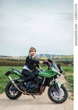 Woman in Leather Jacket on Green Motorcycle. A woman sits confidently on a green motorcycle parked 120651141
