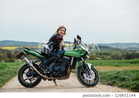 A woman in a leather jacket poses on a green motorcycle parked on a dirt road. The scene conveys a 120651144